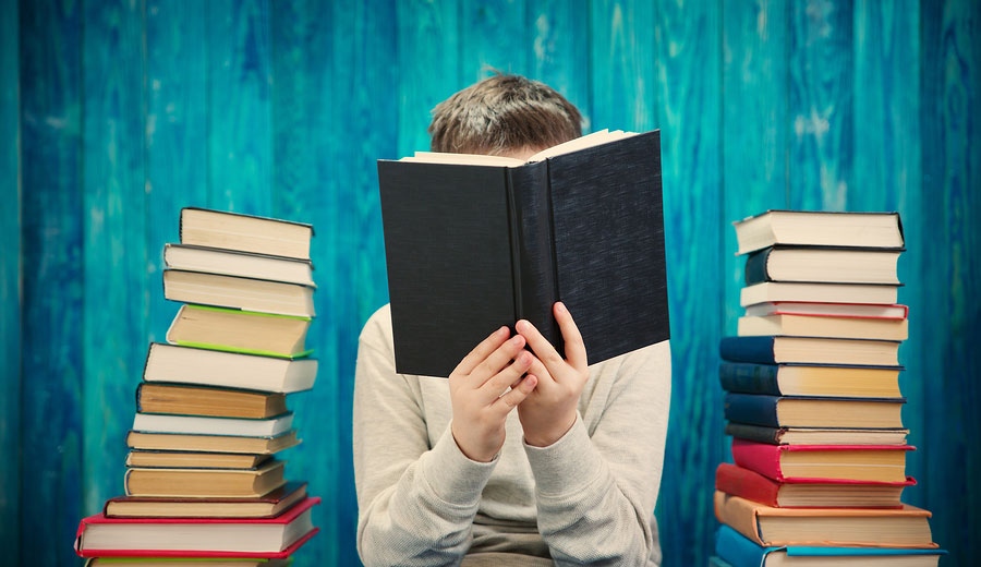 Eight year old child reading a book at home. Boy studying at table on blue background.